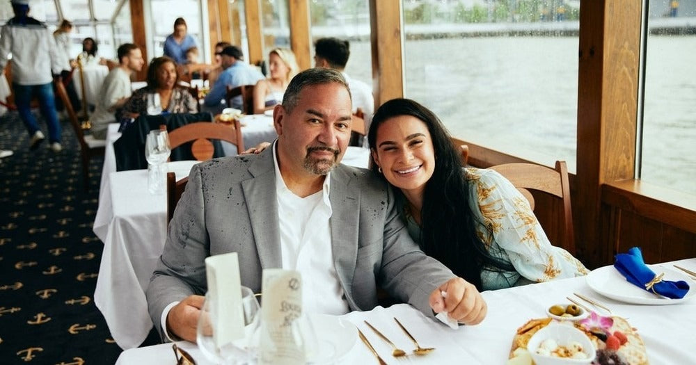 A man and a woman sit closely together, smiling at a table set with silverware and dishes, on an elegant VIP: NYC Dinner Cruise by NYC Water Cruises Inc. with large windows overlooking the water. Other diners are visible in the background, and natural light fills the space.