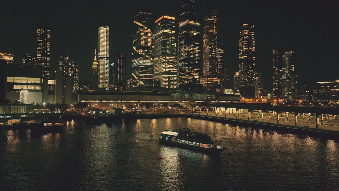  Peaceful night cruise down the Hudson River on the Louisa of the Seas Vessel. The boat glides gently on the water, offering a stunning panorama of New York City's illuminated skyline.  In the distance, iconic landmarks like the Empire State Building and One World Trade Center twinkle against the twilight sky.